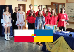 Grupa uczniów znajduje się na szkolnym korytarzu, obok jest stół, a na środku flagi Ukrainy i Polski.